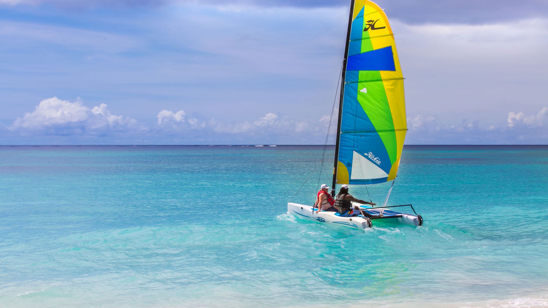 A sailboat with a colorful yellow-blue-green sail carries two people across turquoise ocean under a cloudy sky.