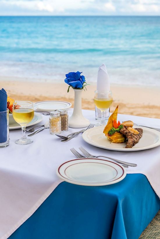 A romantic beachside table set for two with white and blue linens, a plate of food, blue napkin, drinks, and a serene ocean view.